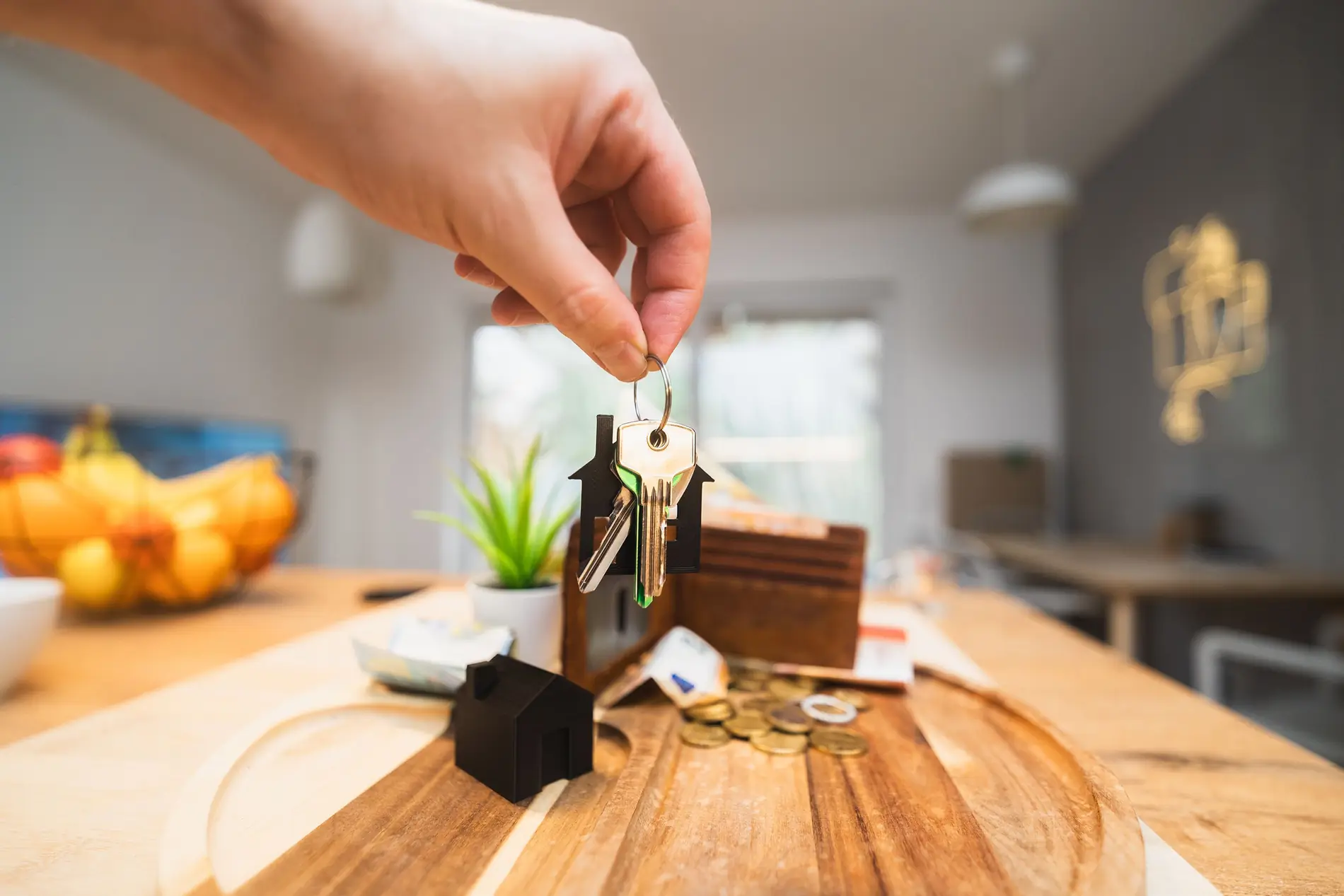 A hand holding a house-shaped keychain over a table with coins and a wallet