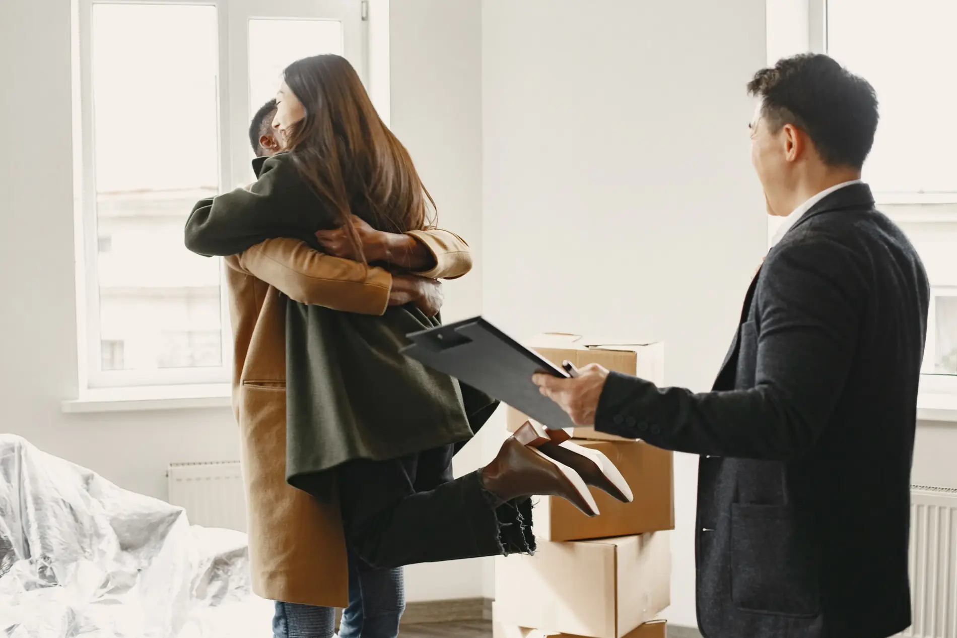A couple embracing while an agent hands over a clipboard beside moving boxes